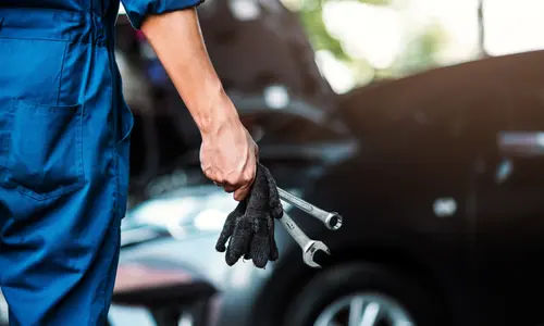 Monteur in een blauwe overall met werkhandschoenen en steeksleutels in de hand, voor een auto in de werkplaats