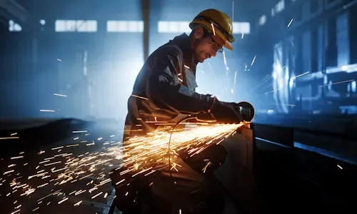 Medewerker met veiligheidshelm slijpt metaal in een fabriekshal, vonken spatten in het rond